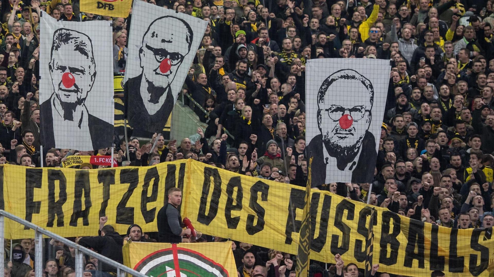 BVB-Fans zeigen Banner mit den Gesichtern von Dietmar Hopp (l-r), Fritz Keller und Karl-Heinz Rummenigge mit roter Clownsnase. Darunter ist ein Banner mit der Aufschrift "Die hässlichen Fratzen des Fußball!". BVB-Fans zeigen Banner mit den Gesichtern von Dietmar Hopp (l-r), Fritz Keller und Karl-Heinz Rummenigge mit roter Clownsnase. Darunter ist ein Banner mit der Aufschrift "Die hässlichen Fratzen des Fußball!".