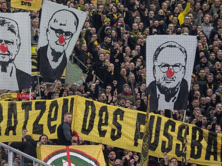 BVB-Fans zeigen Banner mit den Gesichtern von Dietmar Hopp (l-r), Fritz Keller und Karl-Heinz Rummenigge mit roter Clownsnase. Darunter ist ein Banner mit der Aufschrift "Die hässlichen Fratzen des Fußball!".