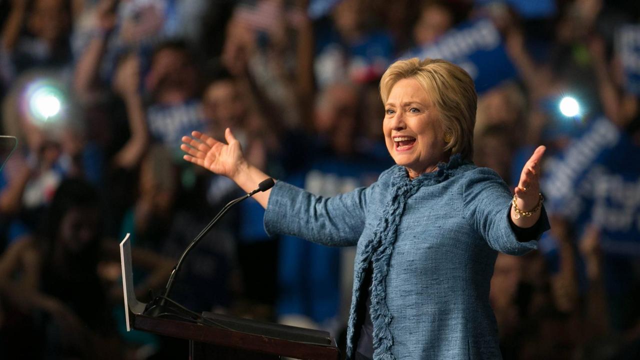 Hillary Clinton vor einem Rednerpult. Sie hat die Arme weit ausgebreitet und strahlt. Hillary Clinton vor einem Rednerpult. Sie hat die Arme weit ausgebreitet und strahlt.