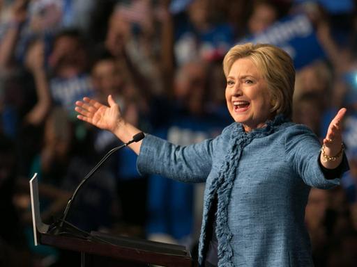 Hillary Clinton vor einem Rednerpult. Sie hat die Arme weit ausgebreitet und strahlt.