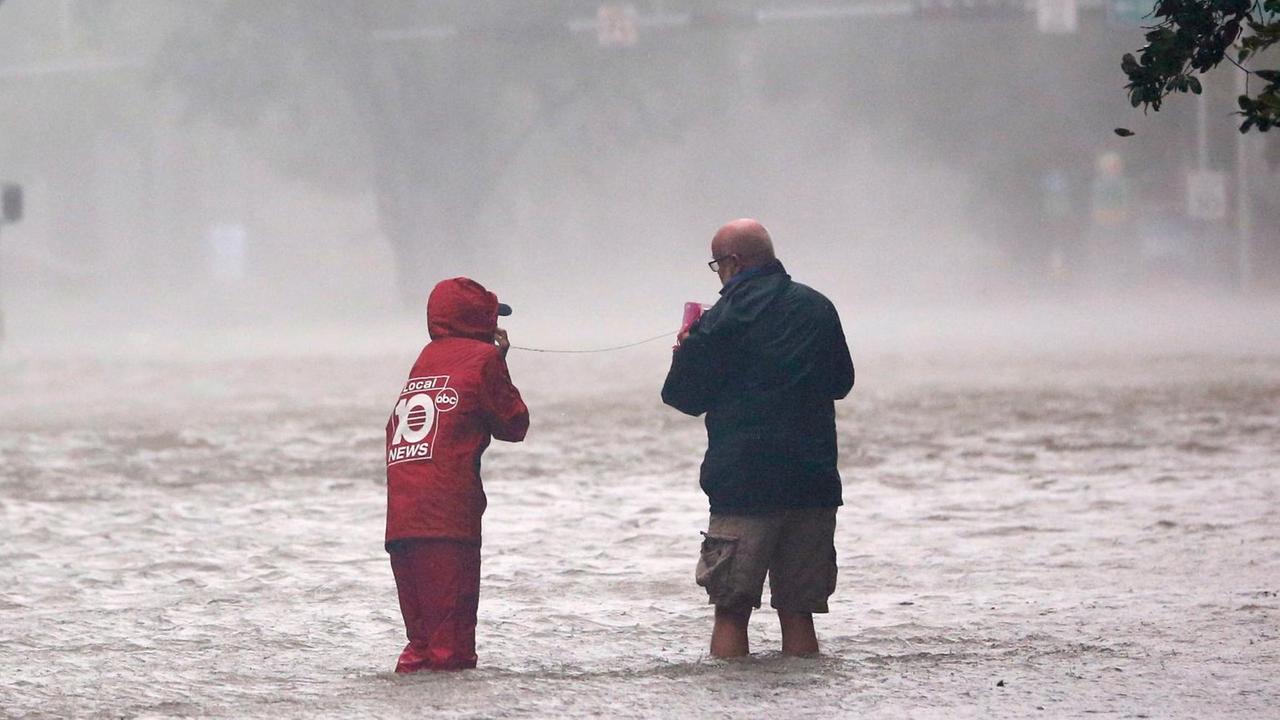 Die beiden stehen im knietiefen und vom Wind bewegten Wasser. Man sieht sie von hinten. Einer hält eine Art Telefon, der andere spricht in einen Hörer. Das Bild ist umrahmt vom den Ästen zweier Bäume. Die beiden stehen im knietiefen und vom Wind bewegten Wasser. Man sieht sie von hinten. Einer hält eine Art Telefon, der andere spricht in einen Hörer. Das Bild ist umrahmt vom den Ästen zweier Bäume.