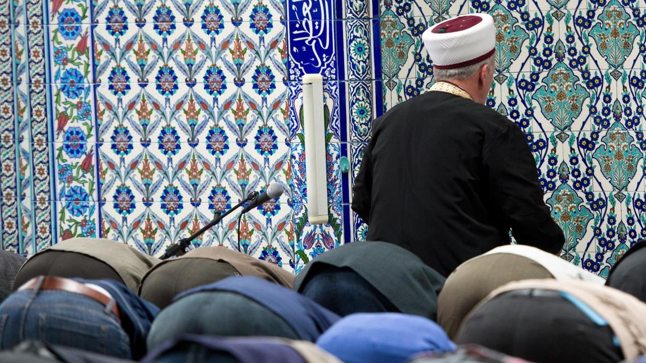 Viele Betende senken den Kopf auf den Boden. Vor ihnen sieht man von hinten den Imam vor einer Wand mit blauen Ornamenten. Viele Betende senken den Kopf auf den Boden. Vor ihnen sieht man von hinten den Imam vor einer Wand mit blauen Ornamenten.