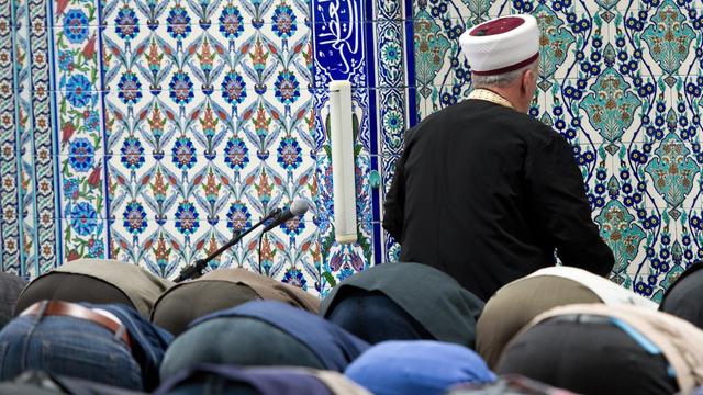 Viele Betende senken den Kopf auf den Boden. Vor ihnen sieht man von hinten den Imam vor einer Wand mit blauen Ornamenten.