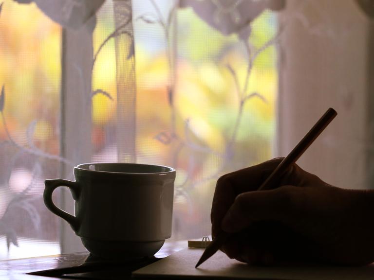 Vor eine, Fenster mit Blick nach draußen steht im Schatten ein Tasse; im Schatten sieht man außerdem eine Hand, die mit einem Stift auf ein Blatt Papier schreibt