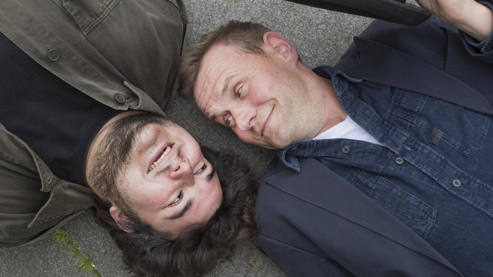Devid Striesow und Axel Ranisch liegen Kopf an Kopf auf dem Boden und schauen sich an.