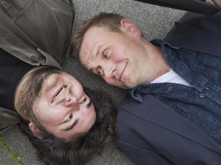 Devid Striesow und Axel Ranisch liegen Kopf an Kopf auf dem Boden und schauen sich an.