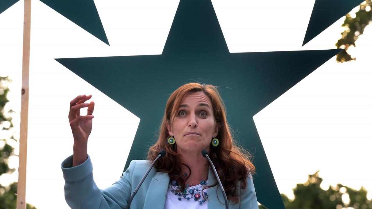 Ein Frau mit langen roten Haaren gestikuliert, vor einem großen grünen Stern stehend, mit der rechten Hand.