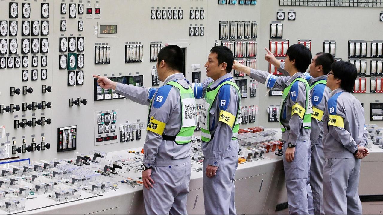 Japanische Techniker in grauen Arbeitsanzügen deuten mit den Fingern auf Anzeigen an einer Wand. Japanische Techniker in grauen Arbeitsanzügen deuten mit den Fingern auf Anzeigen an einer Wand.