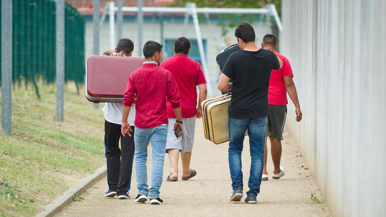 Flüchtlinge laufen am 27.08.2015 mit Koffern bepackt auf einem Weg einer Erstaufnahmeeinrichtung für Flüchtlinge in Ingelheim (Rheinland-Pfalz) entlang.