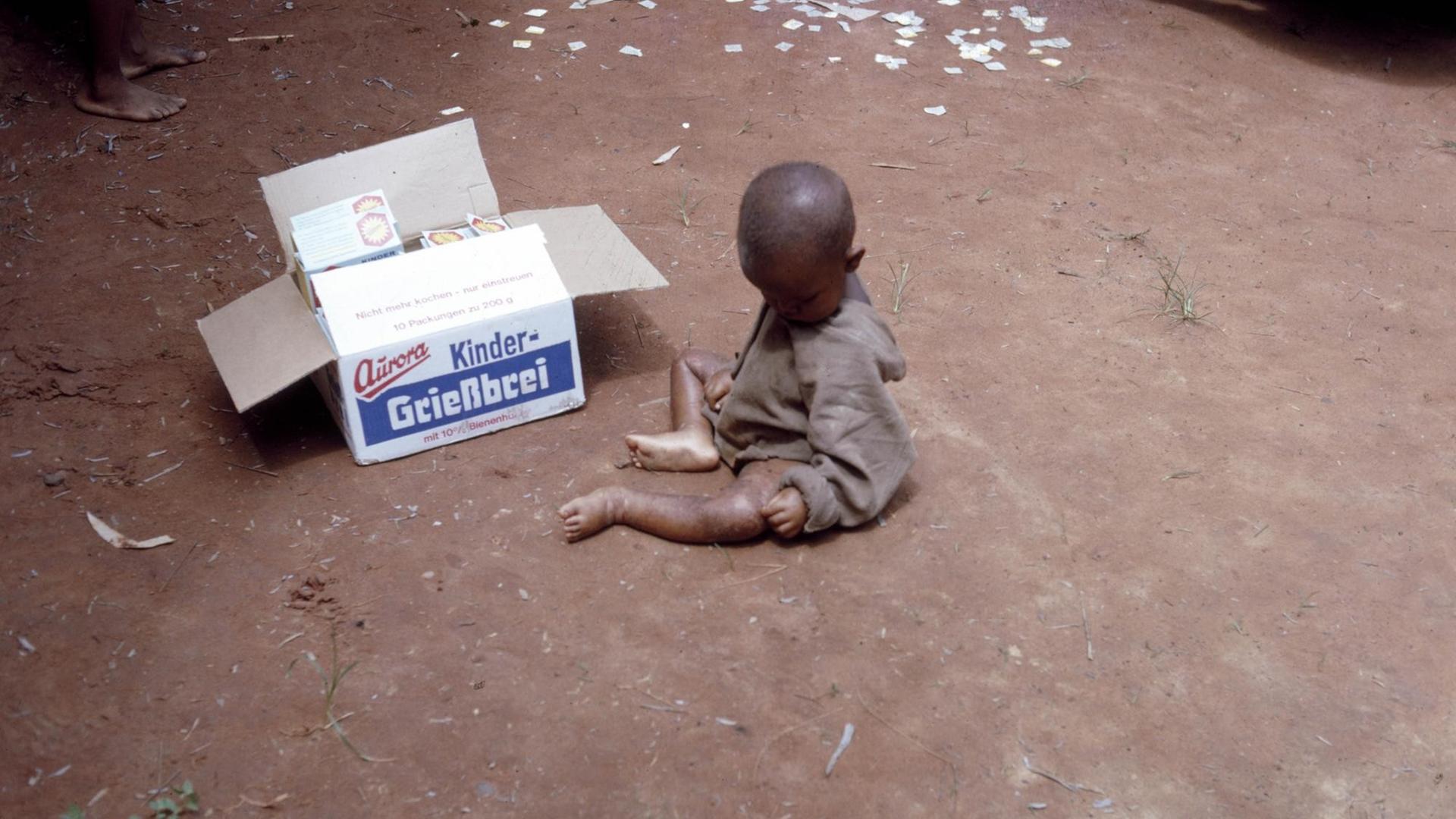 Ein unterernährtes kleines Kind sitzt mit verdecktem Gesicht auf sandigem Boden, daneben eine geöffnete Box, die "Kinder-Grießbrei" der deutschen Marke Aurora beinhaltet. Ein unterernährtes kleines Kind sitzt mit verdecktem Gesicht auf sandigem Boden, daneben eine geöffnete Box, die "Kinder-Grießbrei" der deutschen Marke Aurora beinhaltet.