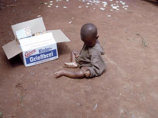 Ein unterernährtes kleines Kind sitzt mit verdecktem Gesicht auf sandigem Boden, daneben eine geöffnete Box, die "Kinder-Grießbrei" der deutschen Marke Aurora beinhaltet.