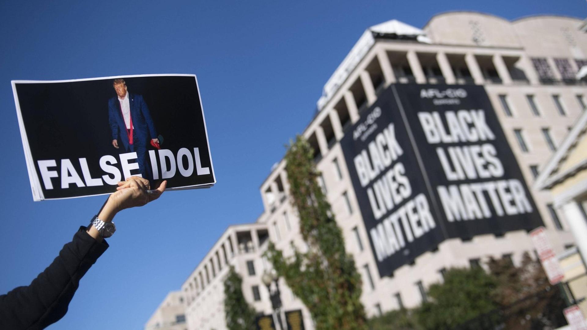 Eine Person hält ein Plakat in die Luft, auf dem neben der Abbildung von Donald Trump der Schriftzug False Idol steht. An einem Gebäude ist ein großes Transparent mit dem Schriftzug Black Lives Matter zu sehen. Eine Person hält ein Plakat in die Luft, auf dem neben der Abbildung von Donald Trump der Schriftzug False Idol steht. An einem Gebäude ist ein großes Transparent mit dem Schriftzug Black Lives Matter zu sehen.