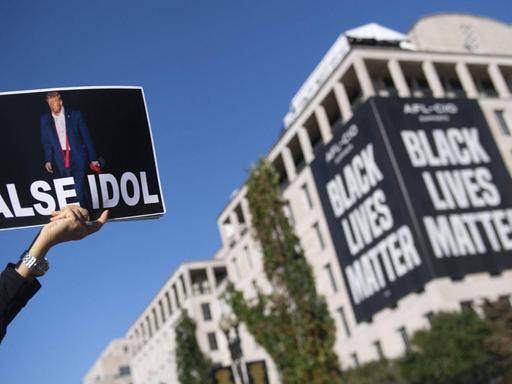 Eine Person hält ein Plakat in die Luft, auf dem neben der Abbildung von Donald Trump der Schriftzug False Idol steht. An einem Gebäude ist ein großes Transparent mit dem Schriftzug Black Lives Matter zu sehen.