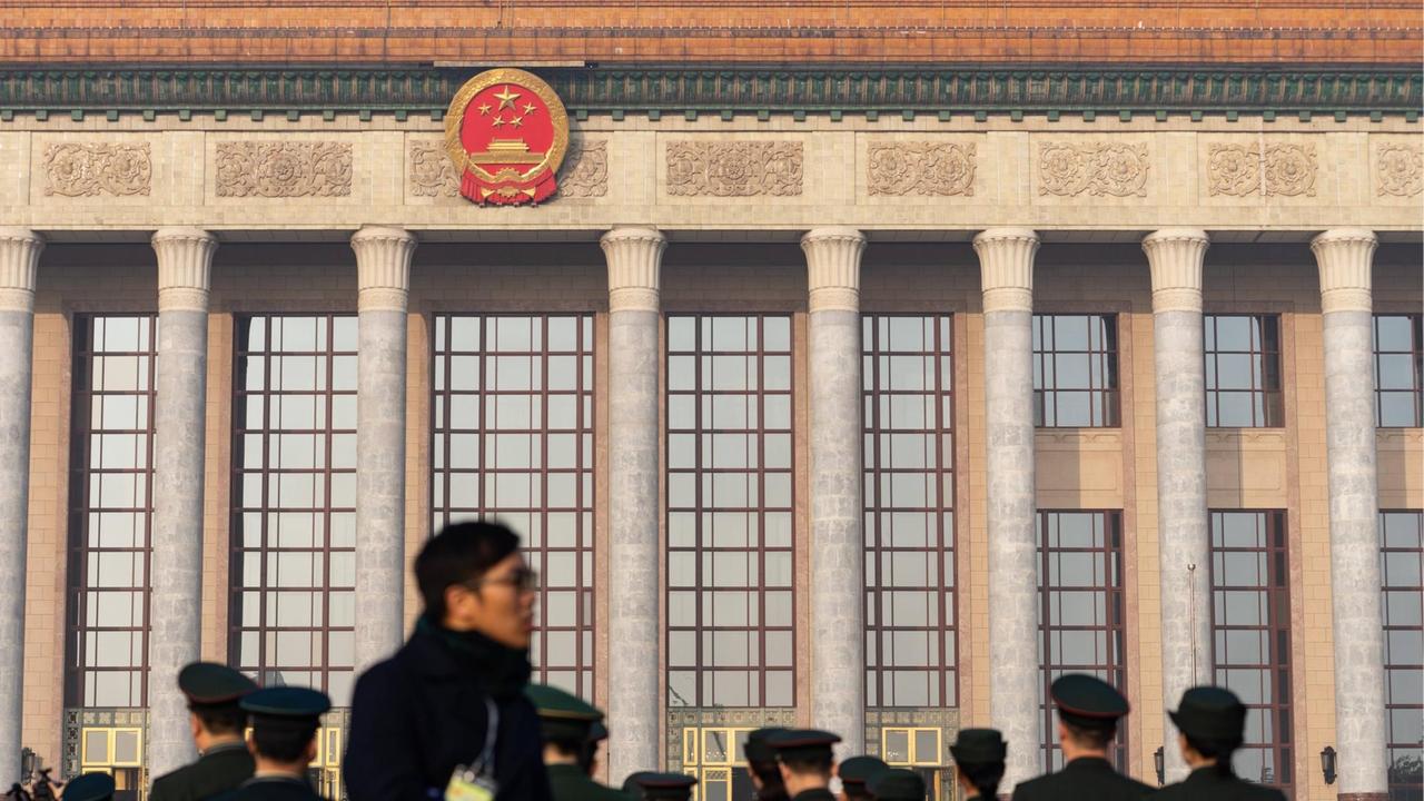 Frontalansicht eines wuchtigen Baus mit kommunistischer Emblematik, davor einige chinesische Besucher.