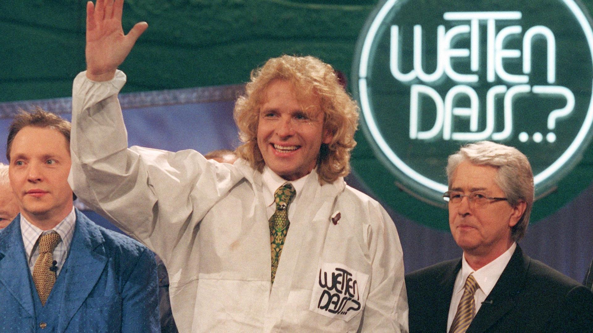 Thomas Gottschalk winkt in hellem Schutzanzug in die Kamera, flankiert von Olli Dittrich und Frank Elstner, im Hintergrund das Logo von "Wetten, dass...?" Thomas Gottschalk winkt in hellem Schutzanzug in die Kamera, flankiert von Olli Dittrich und Frank Elstner, im Hintergrund das Logo von "Wetten, dass...?"