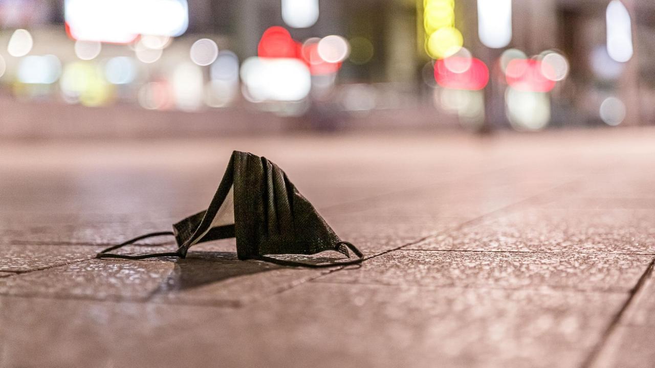 Eine schwarze Mund-Nasen-Bedeckung liegt auf dem Boden in einer Fußgängerzone bei Nacht. Eine schwarze Mund-Nasen-Bedeckung liegt auf dem Boden in einer Fußgängerzone bei Nacht.