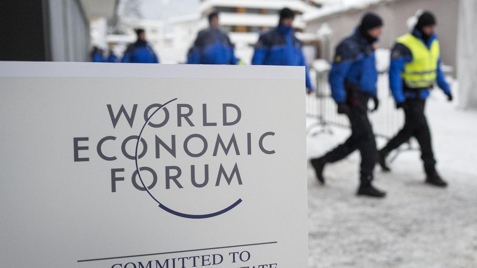 Hinter dem weißen Schild mit der Aufschrift "World Economic Forum - Committed to Improving the State of the World" sieht man unscharf eine Reihe Polizisten durch den Schnee laufen. Hinter dem weißen Schild mit der Aufschrift "World Economic Forum - Committed to Improving the State of the World" sieht man unscharf eine Reihe Polizisten durch den Schnee laufen.
