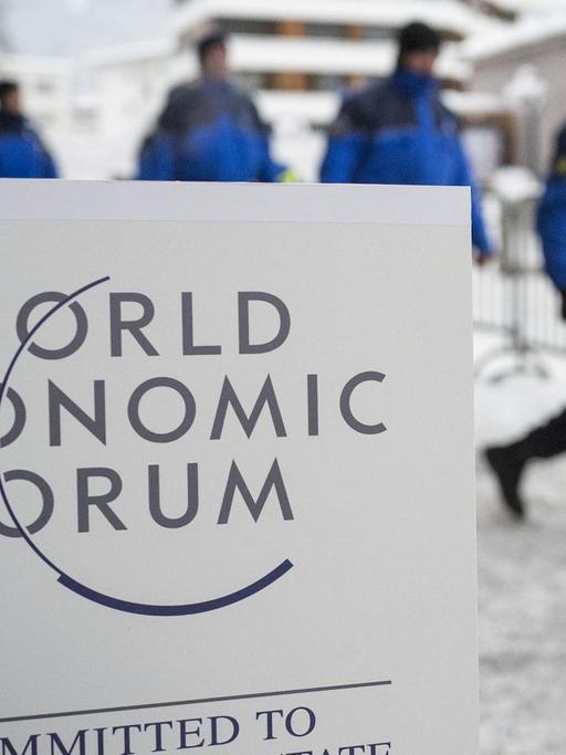 Hinter dem weißen Schild mit der Aufschrift "World Economic Forum - Committed to Improving the State of the World" sieht man unscharf eine Reihe Polizisten durch den Schnee laufen.