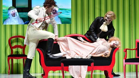 Das Bild zeigt eine Szene aus der Oper "Diodati. Unendlich". Auf einem Sofa liegt eine Frau in Kostüm, zwei Männer stehen neben ihr.
