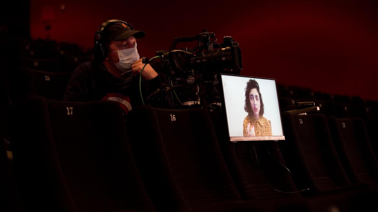Ein Kameramann mit einer Kamera in einem leeren Kinosaal. Auf einem Sitz steht ein Monitor, auf dem ein Dokumentarfilm gezeigt wird. Ein Kameramann mit einer Kamera in einem leeren Kinosaal. Auf einem Sitz steht ein Monitor, auf dem ein Dokumentarfilm gezeigt wird.