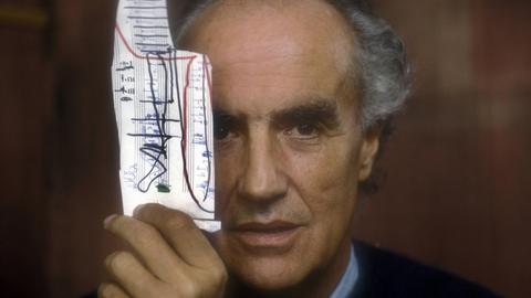 Das Foto zeigt in Frontalansicht den italienischen Komponisten Luigi Nono. Er hält sich ein Stück Notenpapiert vor sein rechtes Auge. Das Foto entstand 1988.