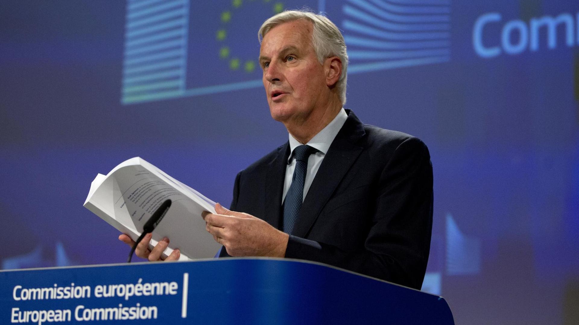 Barnier steht vor einer blauen Wand mit der Aufschrift "European Commission" an einem Pult hinter einem Mikrofon und blättert mit Blick nach vorne in dem Dokument.