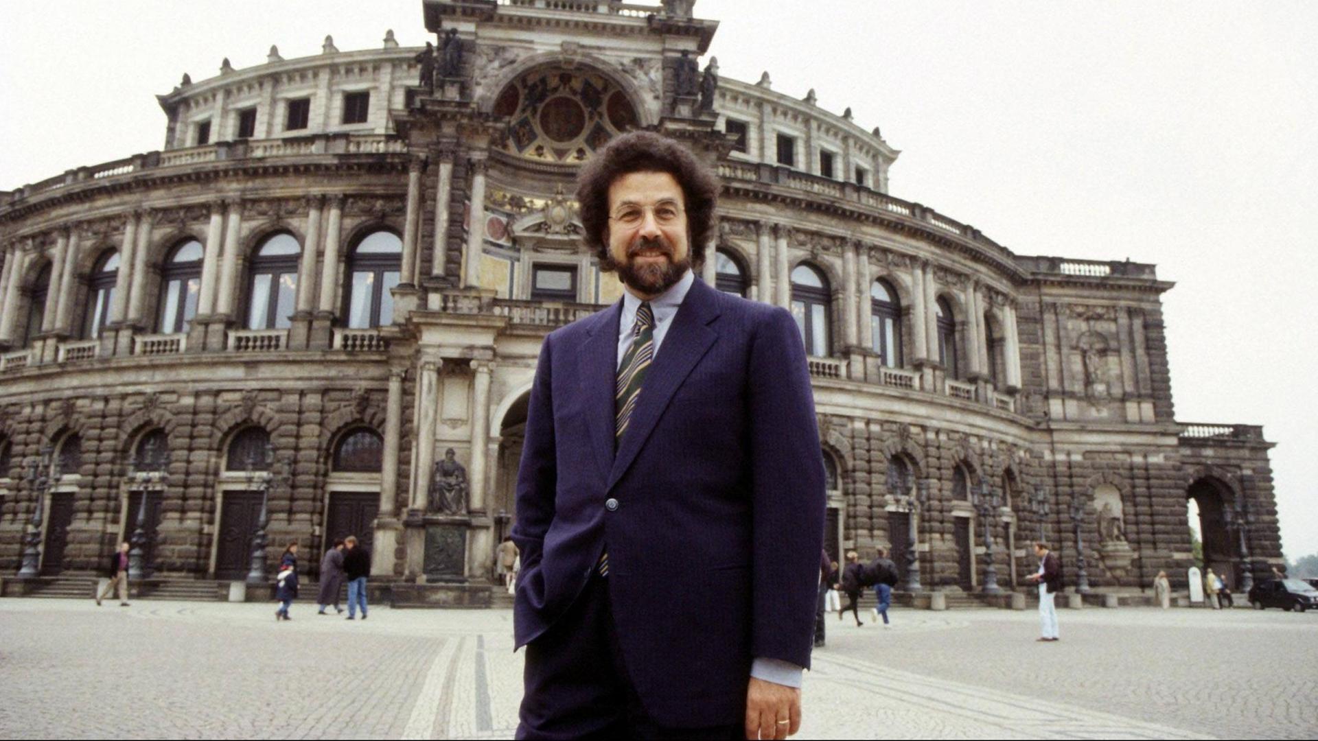 Ein Mann mit dichtem, dunklem Haar und Bart steht lässig vor einem opulenten Bau der Semperoper in Dresden. Ein Mann mit dichtem, dunklem Haar und Bart steht lässig vor einem opulenten Bau der Semperoper in Dresden.