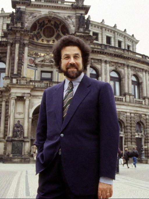 Ein Mann mit dichtem, dunklem Haar und Bart steht lässig vor einem opulenten Bau der Semperoper in Dresden.