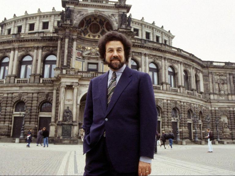 Ein Mann mit dichtem, dunklem Haar und Bart steht lässig vor einem opulenten Bau der Semperoper in Dresden.