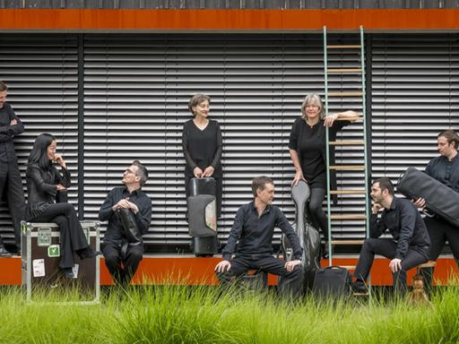 Acht Mitglieder des Ensembles sitzen vor einem Haus mit roten Architekturteilen auf einer Wiese.