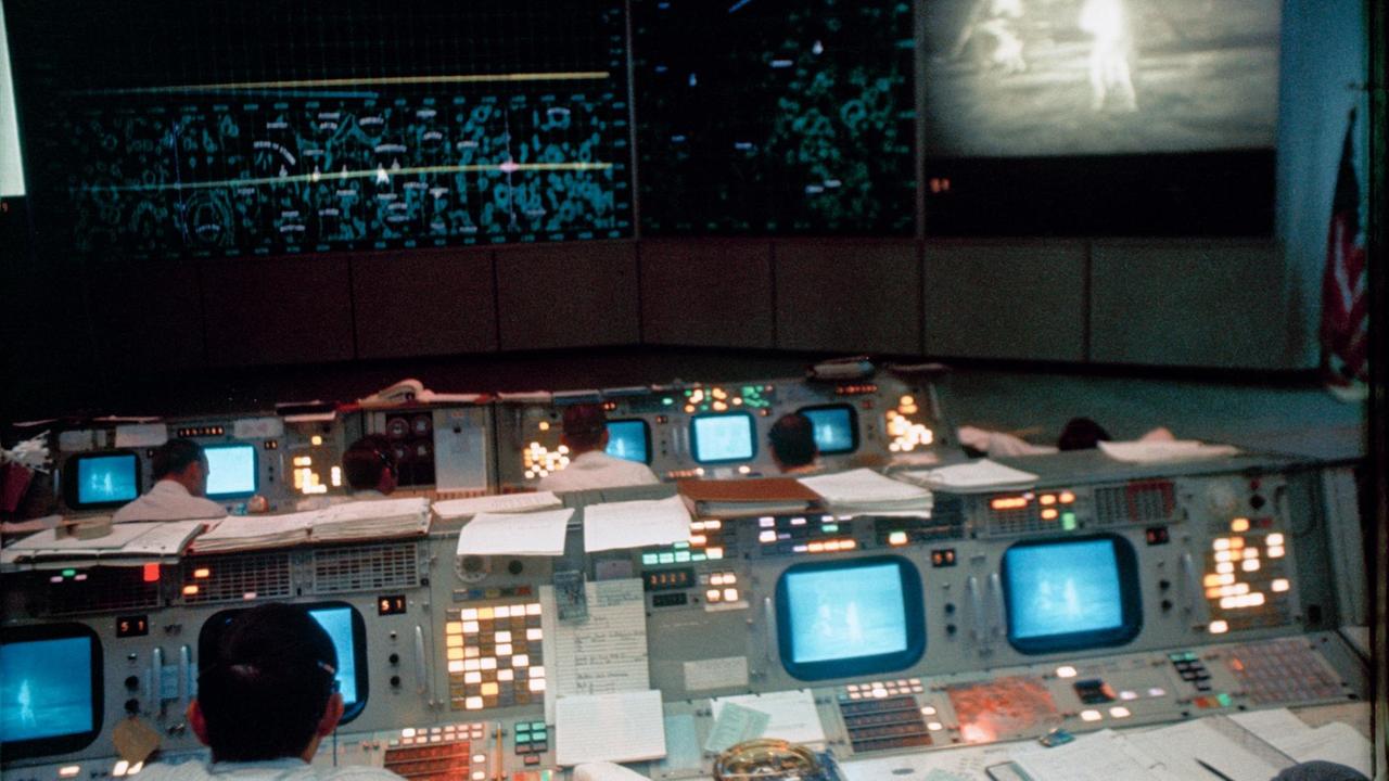 Kontrollcenter der NASA mit Monitoren und Mitarbeitern, an der Wand ein Bildschirm, auf dem die Astronauten Neil. A. Armstrong und Edwin E. Aldrin Jr. auf der Mondoberfläche zu sehen sind.