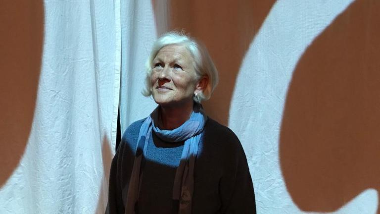 Rita schaut schräg noch oben an der Kamera vorbei und lächelt dabei. Sie hat graue Haare, welche sie seitlich gescheitelt trägt. Ihre Kleidung und Schal sind blau. Rita schaut schräg noch oben an der Kamera vorbei und lächelt dabei. Sie hat graue Haare, welche sie seitlich gescheitelt trägt. Ihre Kleidung und Schal sind blau.