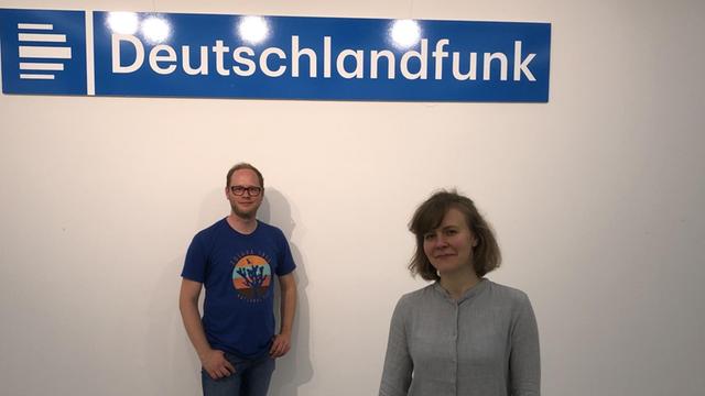 Ein junger Mann im blauen Shirt und eine junge Frau stehen lächelnd vor einer weißen Wand; über ihnen ein Deutschlandfunk-Schild