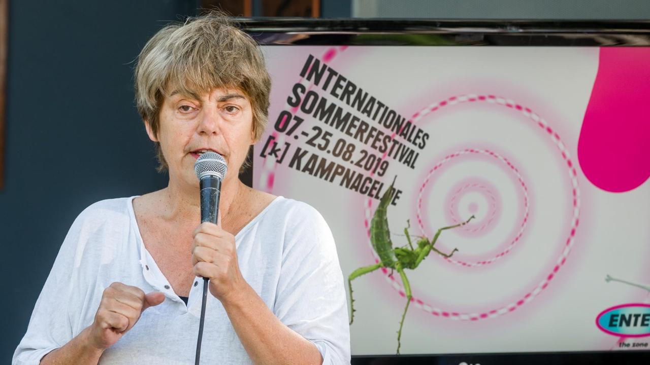 Amelie Deuflhard, Intendantin der Kampnagel-Kulturfabrik ist vor einem Poster des Internationalen Sommerfestivals 2019 mit einem Mikrophon in der Hand zu sehen. Amelie Deuflhard, Intendantin der Kampnagel-Kulturfabrik ist vor einem Poster des Internationalen Sommerfestivals 2019 mit einem Mikrophon in der Hand zu sehen.