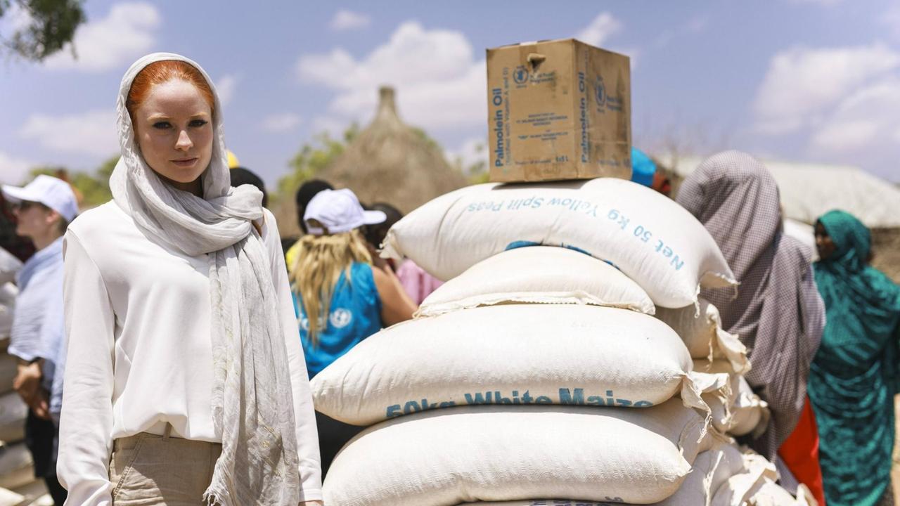 Eine weiße Frau mit roten Haaren steht neben einem stapel Maissäcken. Hinter ihr sind afrikanische Frauen mit Kopftuch. Eine weiße Frau mit roten Haaren steht neben einem stapel Maissäcken. Hinter ihr sind afrikanische Frauen mit Kopftuch.