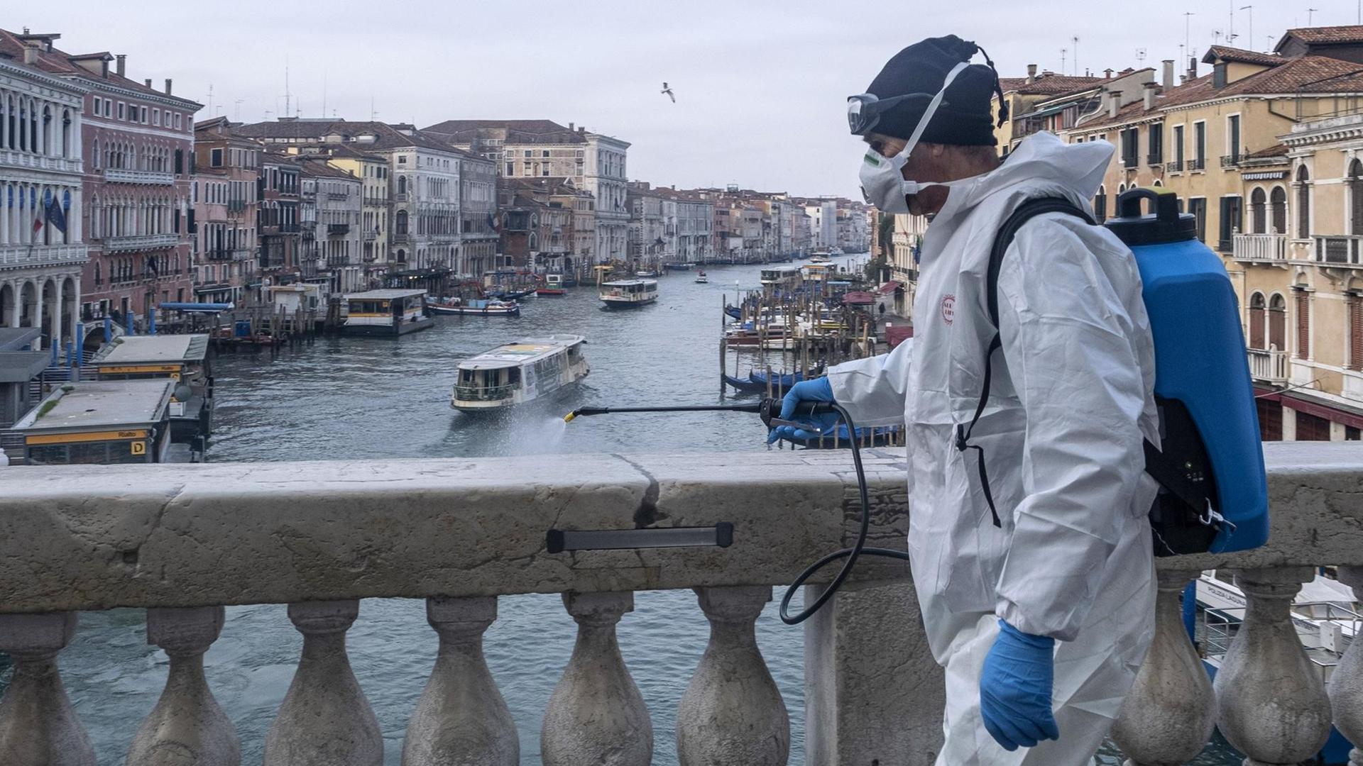 Ein Mann in Schutzanzug desinfiziert den Marmor auf der Rialto-Brücke in Venedig. Im Hintergrund ist der Canal Grande und Häuserfassaden zu sehen. Ein Mann in Schutzanzug desinfiziert den Marmor auf der Rialto-Brücke in Venedig. Im Hintergrund ist der Canal Grande und Häuserfassaden zu sehen.