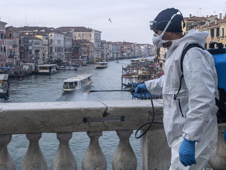 Ein Mann in Schutzanzug desinfiziert den Marmor auf der Rialto-Brücke in Venedig. Im Hintergrund ist der Canal Grande und Häuserfassaden zu sehen.
