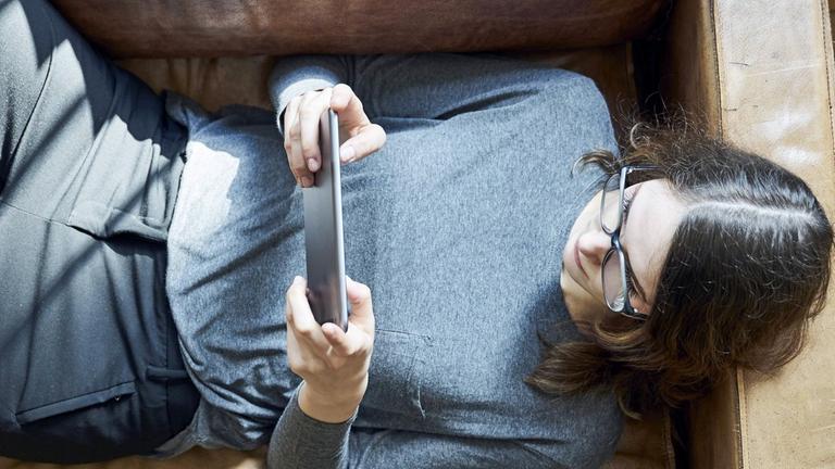 Eine Frau liegt auf dem Sofa und schaut auf den Bildschirm ihres Tablets.