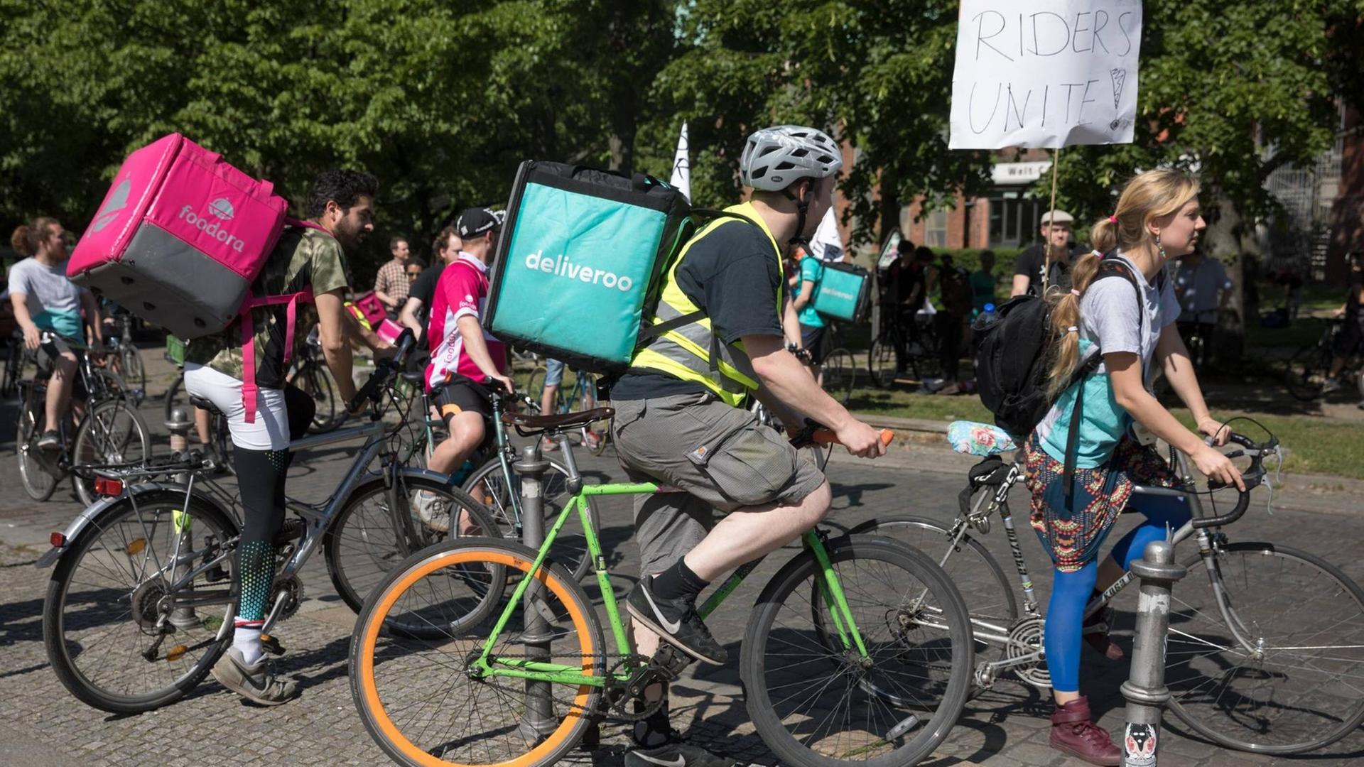 Radfahrer mit pinken und türkisen Liefer-Rucksäcken von Foodora und Deliveroo stehen zusammen an einer Straßenecke. Radfahrer mit pinken und türkisen Liefer-Rucksäcken von Foodora und Deliveroo stehen zusammen an einer Straßenecke.