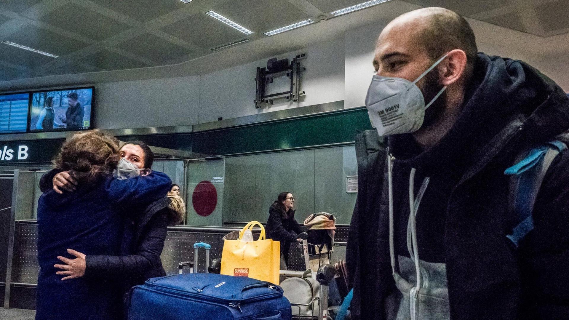 Auf dem Bild sind drei Menschen an einem Flughafen. Ein Mann mit einem Mundschutz steht im Vordergrund. Im Hintergrund stehen zwei weitere Menschen, sie umarmen sich. Sie tragen ebenfalls einen Mundschutz.
