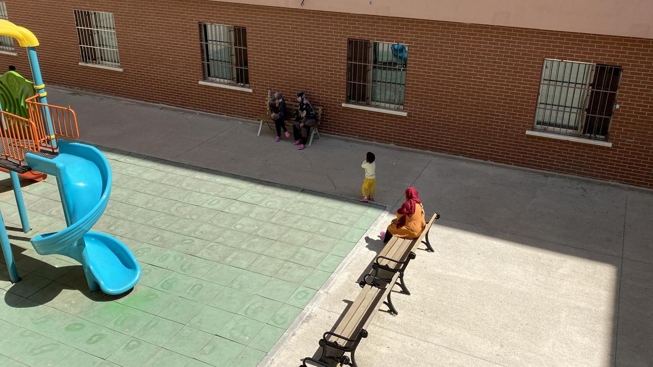 In einem Hof mit vergitterten Fenstern sitzt eine Frau mit Kopftuch und Kind an einer Rutsche. In einem Hof mit vergitterten Fenstern sitzt eine Frau mit Kopftuch und Kind an einer Rutsche.