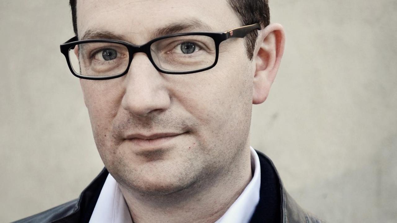 Porträtaufnahme eines Mannes mit westeuropäischem Aussehen in mittlerem Alter, der in die Kamera schaut, eine dunkle viereckige Brille sowie eine schware Jacke mit weißem Hemd trägt. Porträtaufnahme eines Mannes mit westeuropäischem Aussehen in mittlerem Alter, der in die Kamera schaut, eine dunkle viereckige Brille sowie eine schware Jacke mit weißem Hemd trägt.