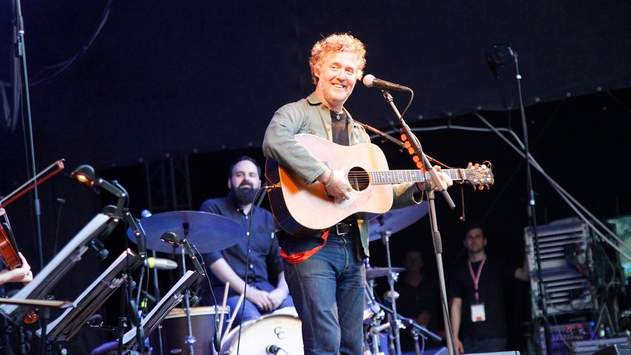 Der Musiker Glen Hansard steht lächelnd, die Gitarre um den Hals auf einer Bühne und spielt Der Musiker Glen Hansard steht lächelnd, die Gitarre um den Hals auf einer Bühne und spielt