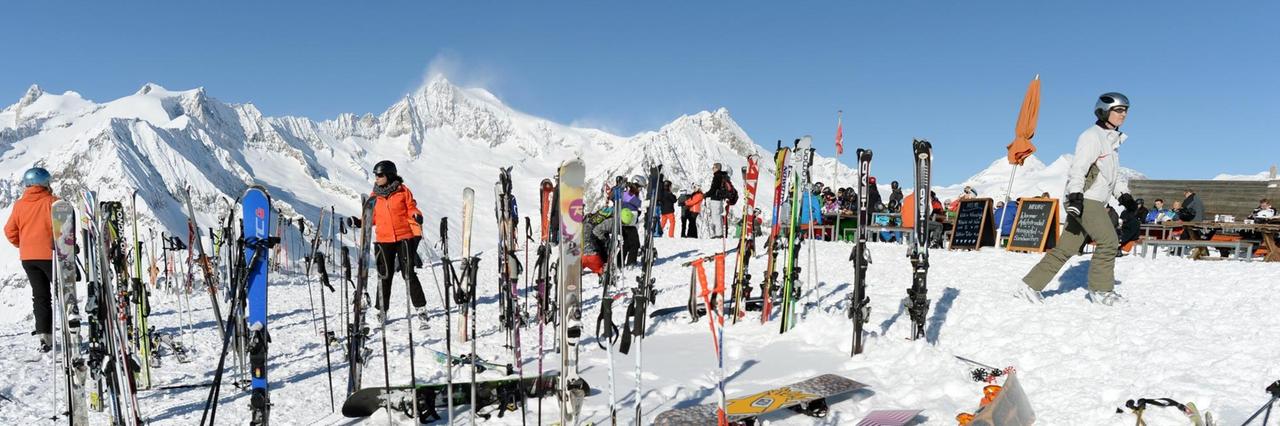 Auf einer schneebedeckten Landschaft stehen mehrere Skifahrer, im Vordergrund liegen Skier und Snowboards. 