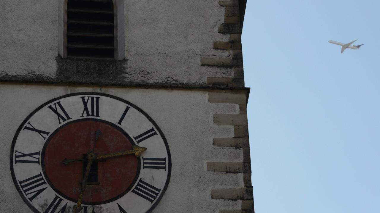 Die Uhr eines Kirchturms in Großaufnahme, daneben weit hinten ein Flugzeug im Landeanflug vor blauem Himmel. Die Uhr eines Kirchturms in Großaufnahme, daneben weit hinten ein Flugzeug im Landeanflug vor blauem Himmel.
