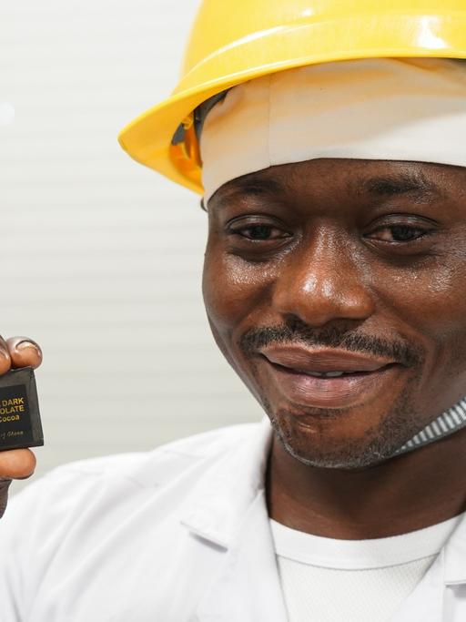 Ein junger Afrikaner mit weißem Kittel und mit gelbem Helm hält ein Stück ghanaische Schokolade in die Kamera.