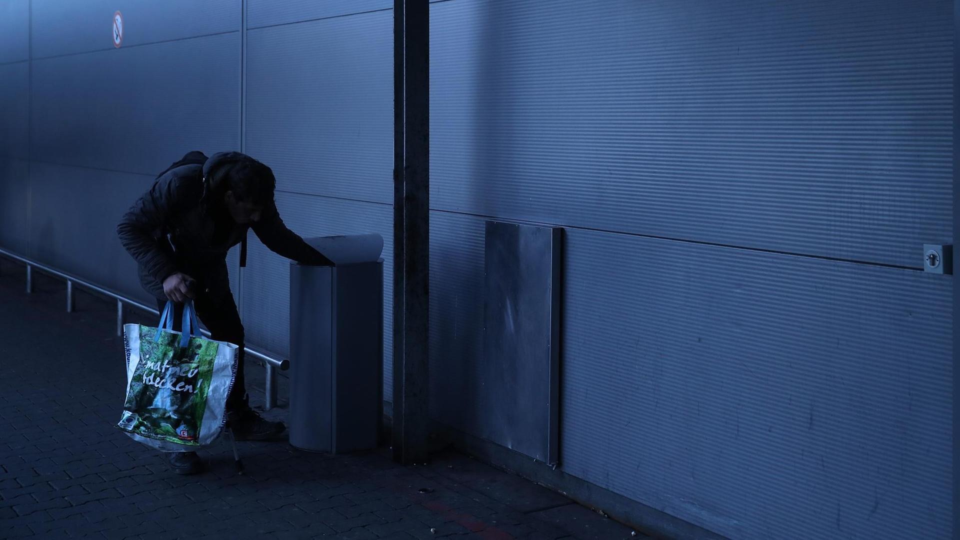 Ein Mann mit einer Plastiktüte lehnt über einem Mülleimer auf der Suche nach Leergut.