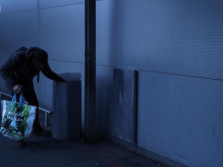 Ein Mann mit einer Plastiktüte lehnt über einem Mülleimer auf der Suche nach Leergut.