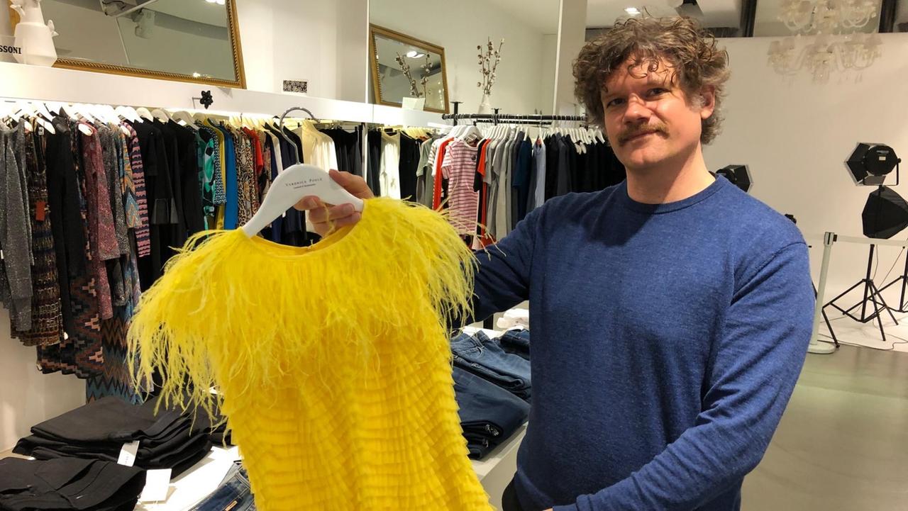 Arik Tremmel hält ein gelbes Kleid auf einem Bügel in der Hand.