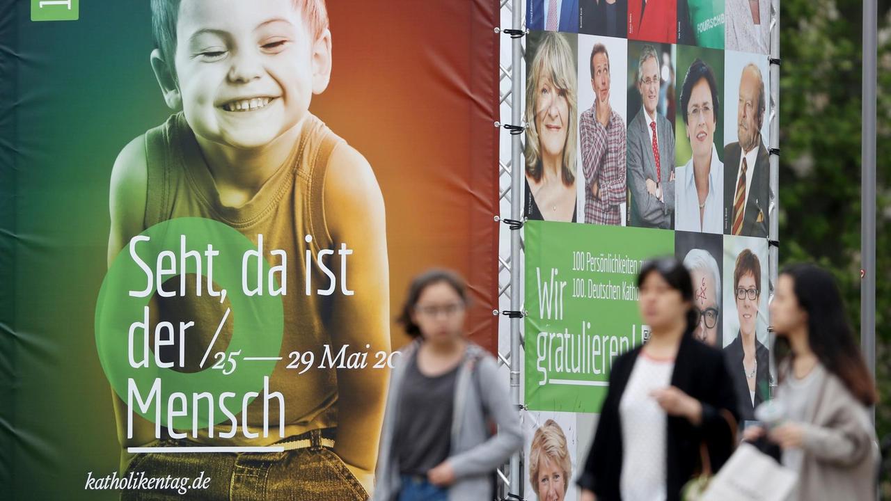 Passanten laufen an einer viereckigen Installation mit Transparenten vorbei. Auf der Vorserseite sieht man das überlebensgroße Bild eines grinsenden Jungen und die Schrift "Seht, da ist der Mensch". Passanten laufen an einer viereckigen Installation mit Transparenten vorbei. Auf der Vorserseite sieht man das überlebensgroße Bild eines grinsenden Jungen und die Schrift "Seht, da ist der Mensch".